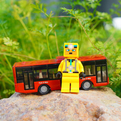 Naklejka premium Lego figure stands in front of a red toy bus on a rock with green foliage