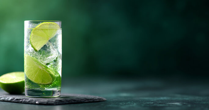 Refreshing glass of sparkling water with lime slices and ice cubes on dark textured surface with green background