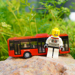 Naklejka premium Astronaut toy figure stands near a red bus on a rock with green foliage background