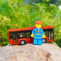 Naklejka premium Lego figure with a red bus on a rock in front of green foliage