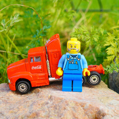 Naklejka premium Lego figure with a red Coca-Cola truck on a rock with green foliage background