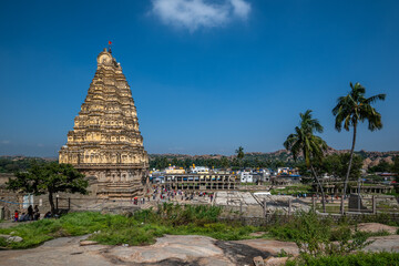Hampi, India - 01 November 2025: View of the towering Virupaksha Temple bathed in warm sunlight, contrasting against the clear blue sky and lush greenery.