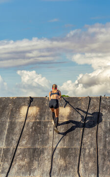 Rear view of a female athlete in short, tight-fitting athletic wear, climbing and reaching the top of a wooden obstacle wall with ropes against a bright, cloudy sky.