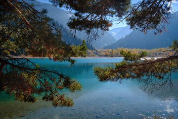 Issyk Lake in autumn. The crystal clear water reflects the sky and the mountains, and the golden trees on the shore create a picturesque frame for this mountain landscape.