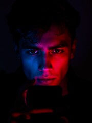 Intense gaze of a young man illuminated by vibrant red and blue neon lights, captivated by his smartphone screen in the darkness