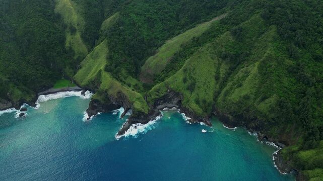 Advancing aerial of Dingalan, Aurora, pushing forward over lush green cliffs and rugged coves as turquoise waters crash against the shoreline under a partly cloudy sky.