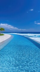 Obraz premium Sparkling blue luxury resort infinity pool merges with ocean waves under a clear blue sky with white clouds and a sandy beach shoreline with a small green tree.