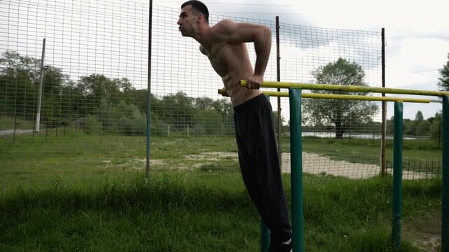 Young sportsman doing push ups on parallel bars at nature. Strong sporty man exercising at sports ground. Male shirtless athlete training hard outdoor. Concept of active sportive lifestyle. Slow mo