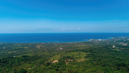 Fototapeta premium Aerial perspective of a vibrant green landscape, seaside village, and vast blue ocean beneath a clear sky, emphasizing the tranquility of nature