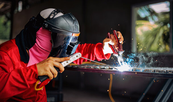 Welder wearing a red long-sleeved shirt and a protective mask is welding a steel grating, Industrial Welder With Torch - Powered by Adobe
