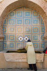 Operating old wall fountain in Tetouan, Morocco, covered with vibrant Islamic tiles and Arabic geometric patterns, with a local woman collecting water.