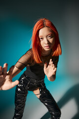 asian woman with piercing and red dyed hair gesturing and looking at camera on blue background