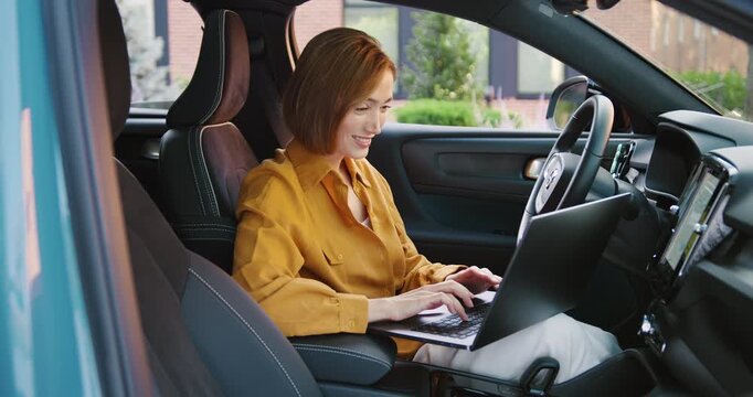 Smiling Caucasian woman working remotely on a laptop while sitting in her parked car near a modern office building, focused on her task.