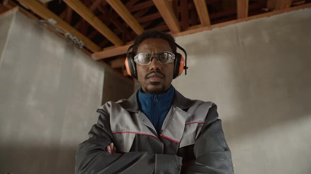 Zoom-in portrait of Black male construction worker wearing safety glasses and earmuffs posing confidently with folded arms at unfinished construction site
