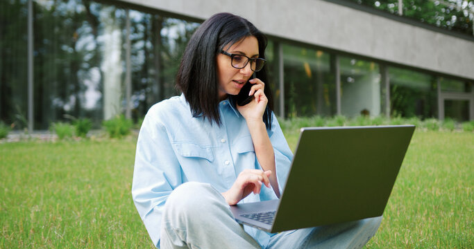 Beautiful young woman sitting in a city park, talking on the phone while typing on a laptop. Concept of online education, IT, business and freelance work in an urban outdoor setting