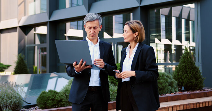 Two business businesspeople using laptop and smartphone Caucasian man businessman guy male talking discuss arguing blond woman businesswoman female girl in city.