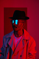 Half-turned portrait of African American male model in fashionable trenchcoat, sunglasses and fedora posing in neon lighting