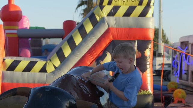 Bull ride captures pure childhood excitement as young boy holds on tight, laughing with joy under bright sunset. Bull movement, inflatable playground and happy energy. cheerful boy enjoys bull ride.