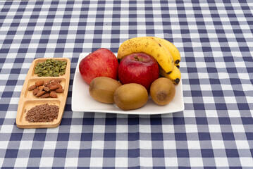 Platter of fruit (apples, kiwis, bananas) and a divided tray with almonds, pumpkin seeds, and flax seeds on a navy blue and white plaid tablecloth. Ideal for healthy snack and diet concepts.