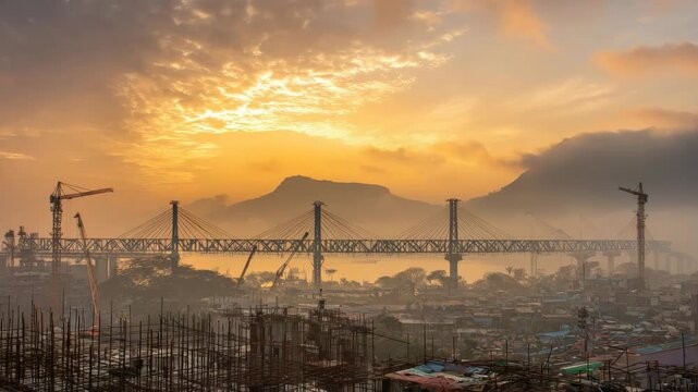 Suspension Bridge Over Dense Cityscape at Warm Sunset with Rolling Hills and Construction Cranes in Landscape Format