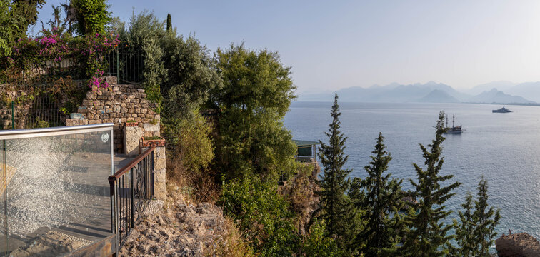 Panoramic view from Kalei&ccedil;i viewpoint with glass railing and old stone wall, overlooking sea with yachts and Taurus Mountains in haze. Antalya, Turkey.

