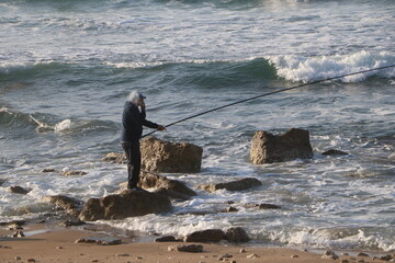 Man fishing on the rocky shore of the sea at sunset. Peaceful seascape scene symbolizing patience, solitude, and connection with nature.