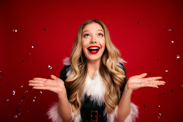 Joyful woman in festive Santa style dress celebrates Christmas joy with confetti against a bold red...