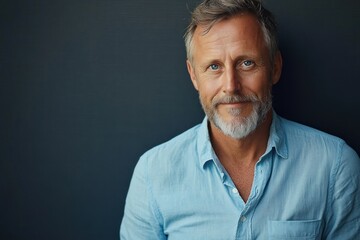 Fototapeta premium Smiling middle-aged man with grey beard and short hair wearing a light blue shirt against a dark background, conveying warmth and confidence