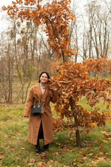 Young attractive woman wearing a brown coat standing under an oak tree in autumn park