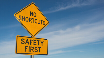 A yellow no shortcuts sign above a safety first sign against a blue cloudy sky during the daytime