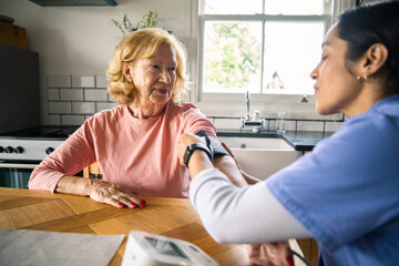 Senior woman smiling as adult nurse checks blood pressure in kitchen