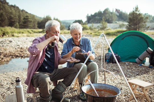 Senior couple smiling over campfire stew at riverside campsite