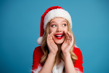 Joyful Santa girl in red dress and fluffy hat smiles with hands near her mouth against blue...