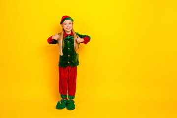Little girl dressed as an elf against a vibrant yellow background showing cheerful thumbs up during the festive season
