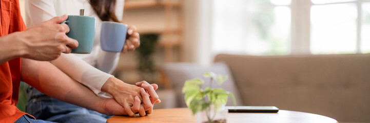 Asian adult couple holding hands drinking coffee tea mugs enjoying cozy home relaxation bonding together living room