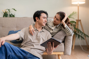 Young Asian couple home relax enjoy digital connection on tablet sharing screen in cozy living room...