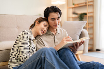 Obraz premium Young Asian couple adults relax home reading book together cozy living room bonding peaceful leisure domestic life