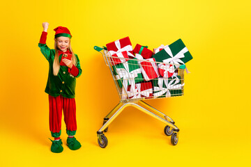 Excited girl dressed as an elf using a phone near a shopping cart full of gifts against a bright yellow background