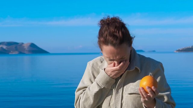 Woman smelling and rejecting an orange. Woman standing by azure sea, rejecting orange with strong facial expression of disgust after smelling fruit, displaying clear sensory aversion to citrus