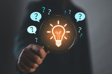 Businessman holding magnifying glass with glowing light bulb and question marks, representing problem solving, finding solutions, critical thinking, and innovative decision making.
