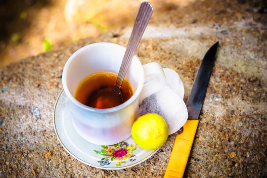 An outdoor still life featuring a hot cup of tea with a spoon, a lemon, two tea bags, and a small knife resting on a floral saucer, all placed on a rough, sun-dappled stone surface - Powered by Adobe