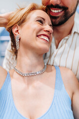 Happy couple shopping for jewelry in a luxury store, with the man lovingly placing a stunning necklace around his smiling blonde girlfriend's neck, celebrating their romance