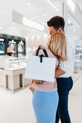 An elegant woman in a blue dress stands next to her partner at the entrance of an exclusive jewelry store and holds a shopping bag.