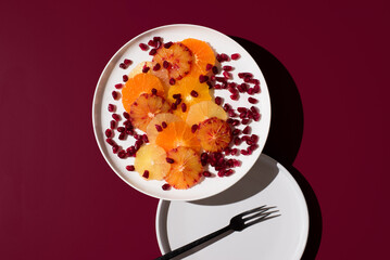 Rodajas de cítricos variados: naranja, limón, mandarina, naranja sanguina, lima y semillas de granada en un plato blanco sobre fondo granate. Vista superior