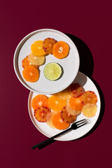 Rodajas de cítricos variados: naranja, limón, mandarina, naranja sanguina, lima en un plato blanco sobre fondo granate. Vista superior
