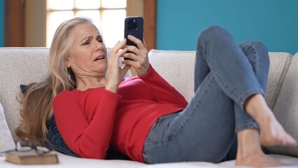Mature woman with blonde hair is lying on a white couch, wearing a red shirt and blue jeans. She is smiling and using a smartphone.