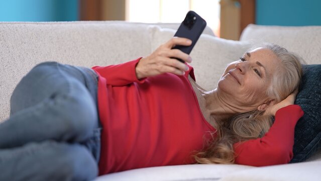 Middle-aged woman lies comfortably on her couch, smiling laughing and waving as she has video chat on smartphone. Natural light fills the cozy living room, creating a warm atmosphere.