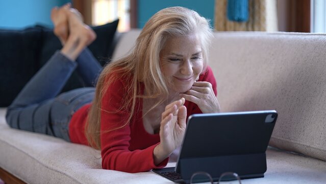 A mature middle age woman with long blonde hair smiles and waves at the camera of a tablet while lying on a white couch.