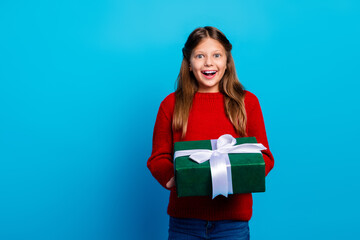 Little girl in red sweater presents a green gift box with a white ribbon against blue background...
