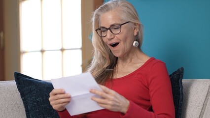 Happy middle-aged woman with long, blonde hair reads good news letter, with concern. She sits comfortably on a couch with a blue wall background.